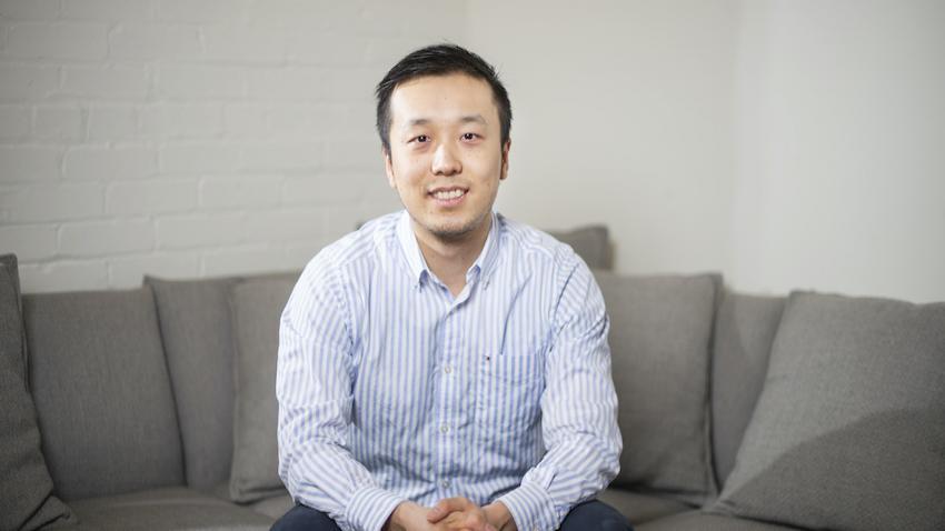 Henry Shi in a blue-and-white striped button-up shirt, sitting on a gray couch