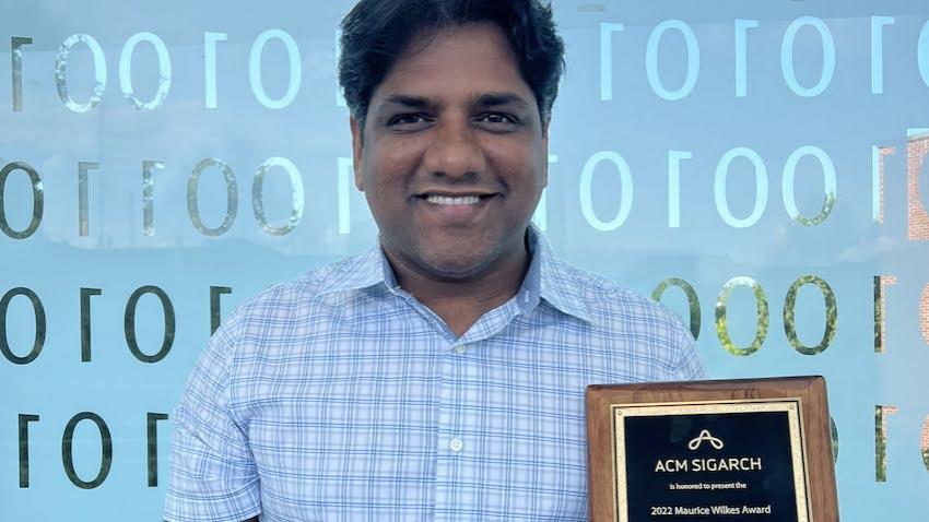 Moin Qureshi standing in front of binary code, holding the 2022 Maurice Wilkes Award plaque