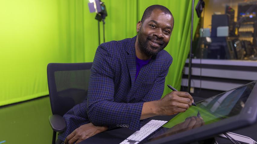 Fisayo Omojokun sitting down, writing on a screen with a stylus, with a greenscreen in the background