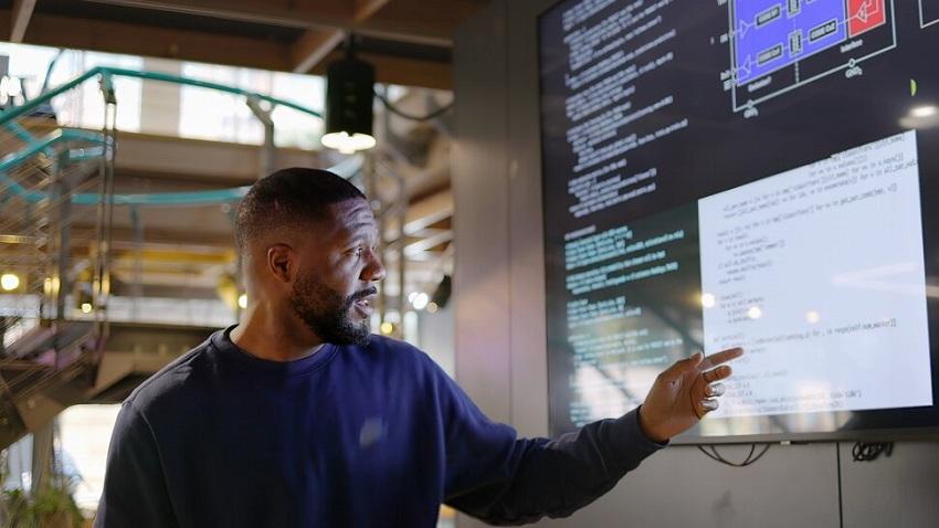 Man in an office pointing at code on a TV-sized monitor