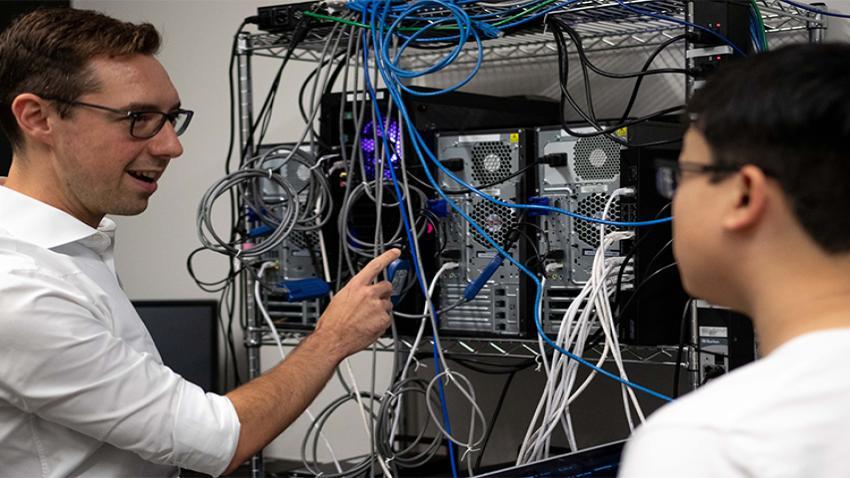 Two people talking in front of a shelf of CPUs and cables, one of them pointing at the hardware