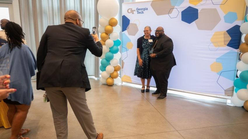 Someone taking a photo of two people standing in front of a Georgia Tech backdrop