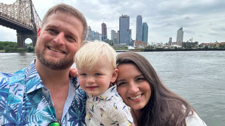 Alex Adams with his wife and young child in front of a bridge over a river