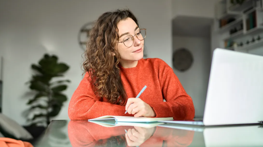 Woman at a desk facing a laptop and writing on a piece of paper
