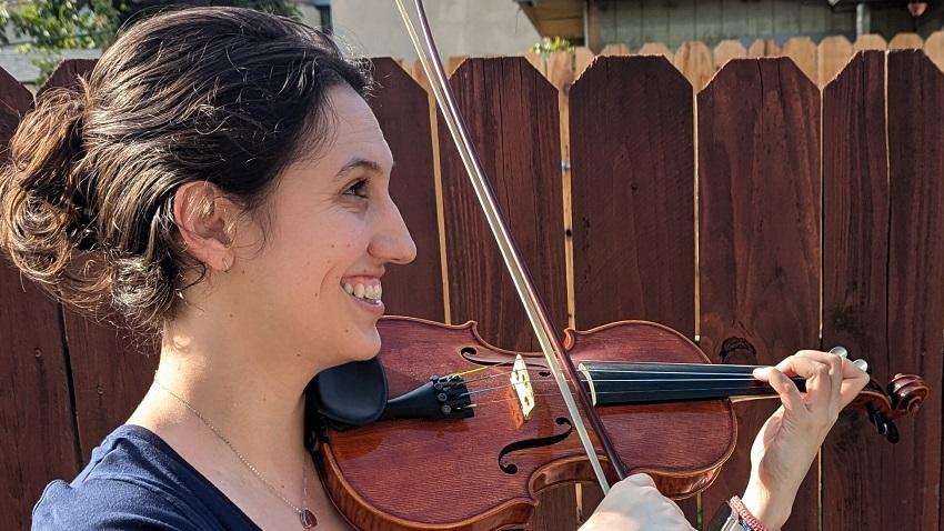 Mercedes Quintana standing outside in front of a brown fence playing violin