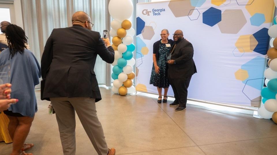 Someone taking a photo of two people standing in front of a Georgia Tech backdrop