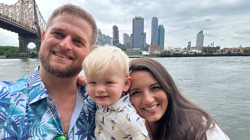 Alex Adams with his wife and young child in front of a bridge over a river